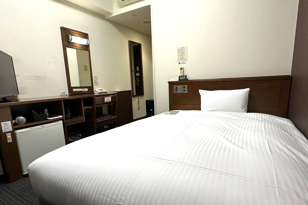 A hotel room with a bed, a desk, a mirror, a small refrigerator, and a nightstand with a clock and a phone.