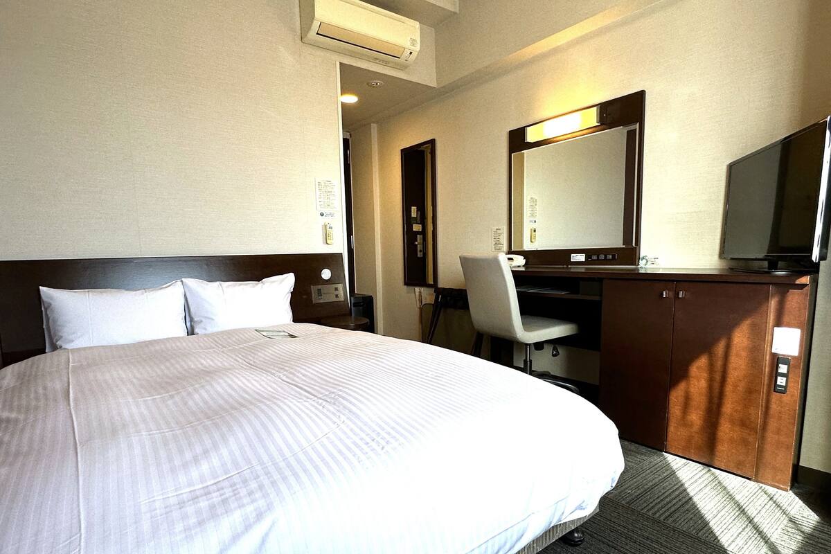 A hotel room with a large bed, a desk with a chair, a mirror, and a television.