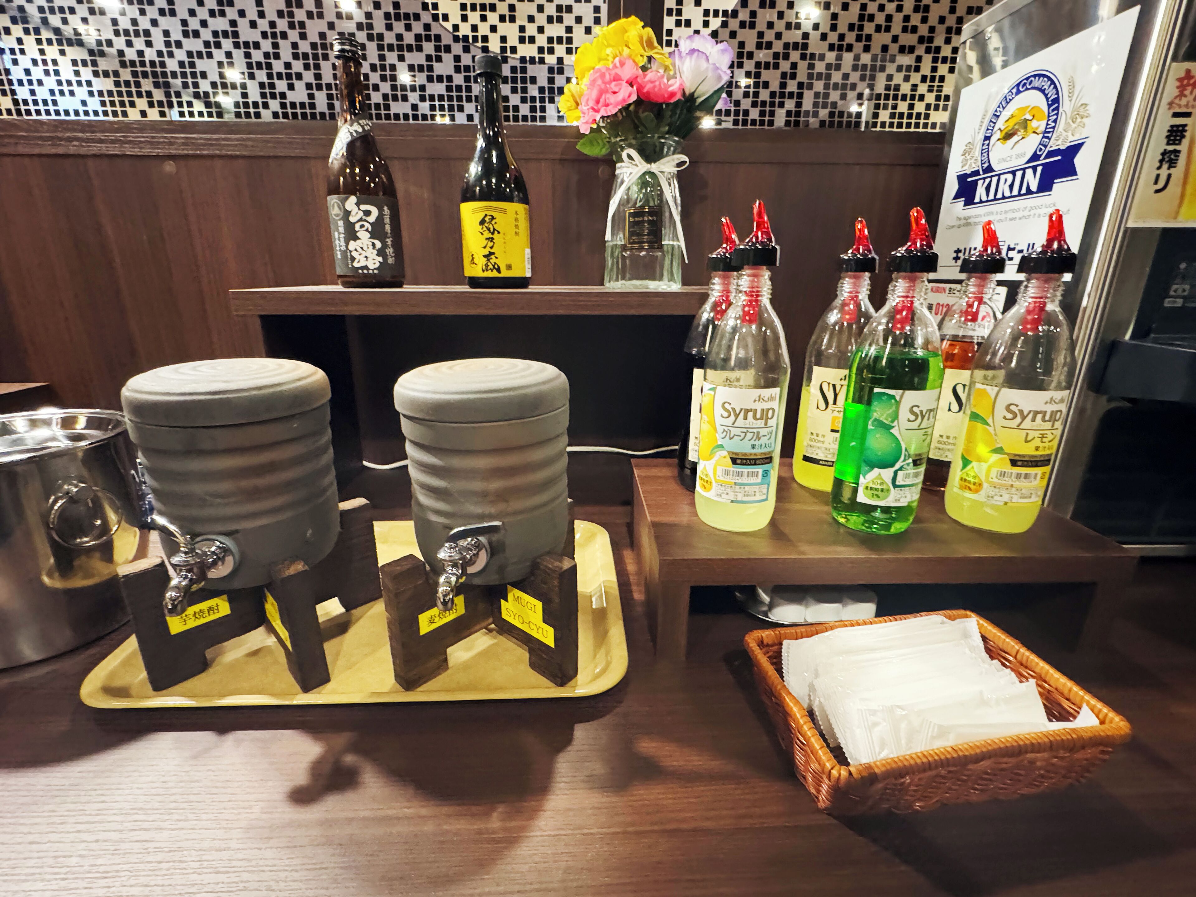 A kitchen counter with various bottles, a syrup dispenser, and a basket of napkins.