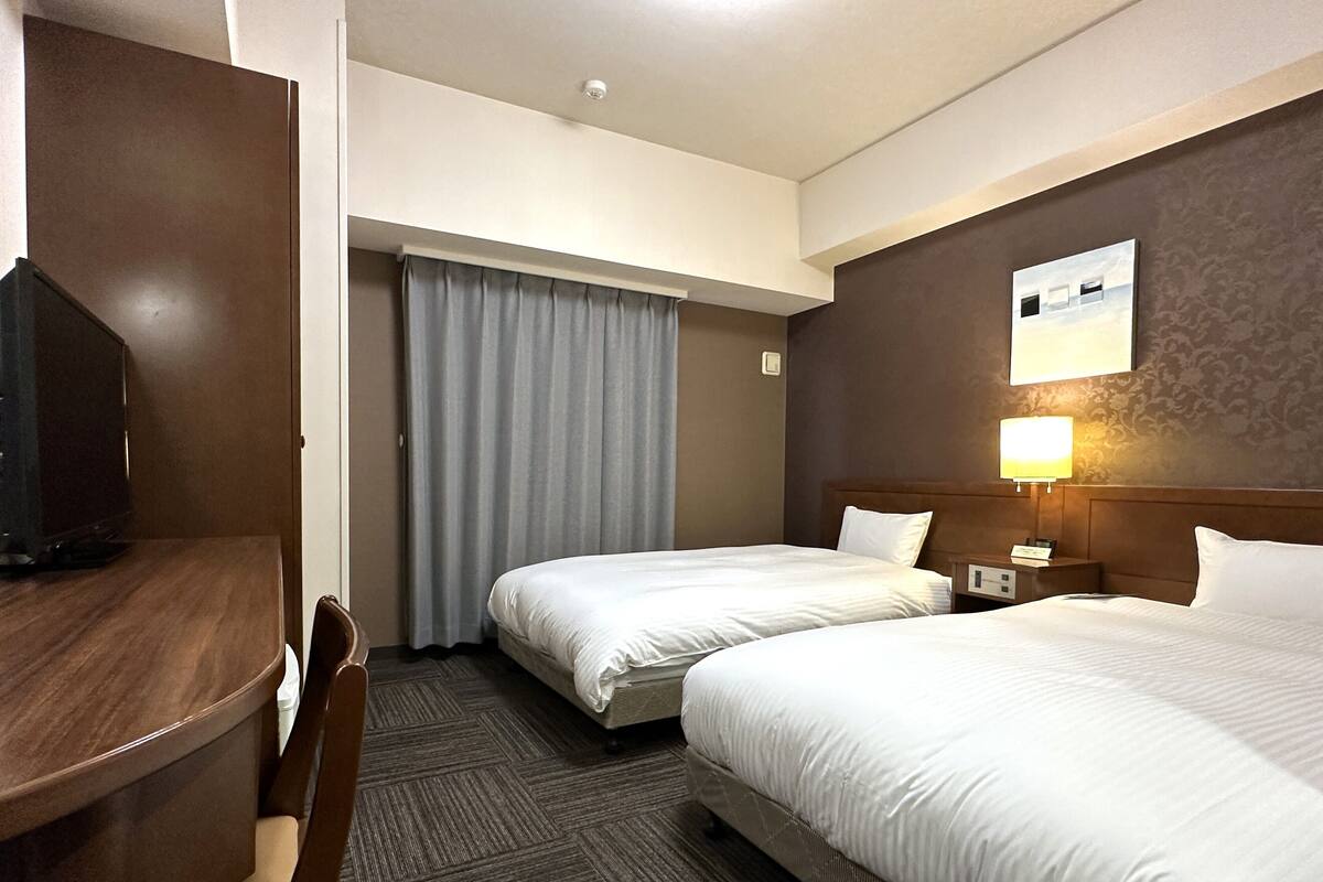 A hotel room with two beds, a desk, a chair, a TV, and a lamp.