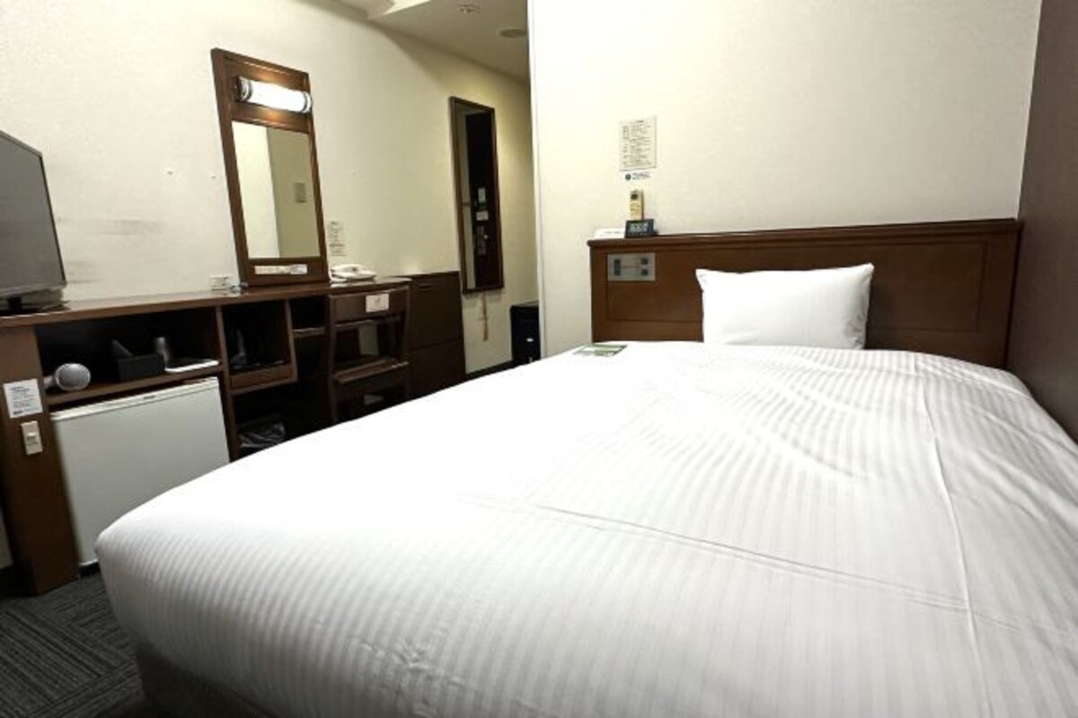 A hotel room with a large bed, a wooden headboard, a desk with a mirror, a small radiator, and a wall-mounted clock.