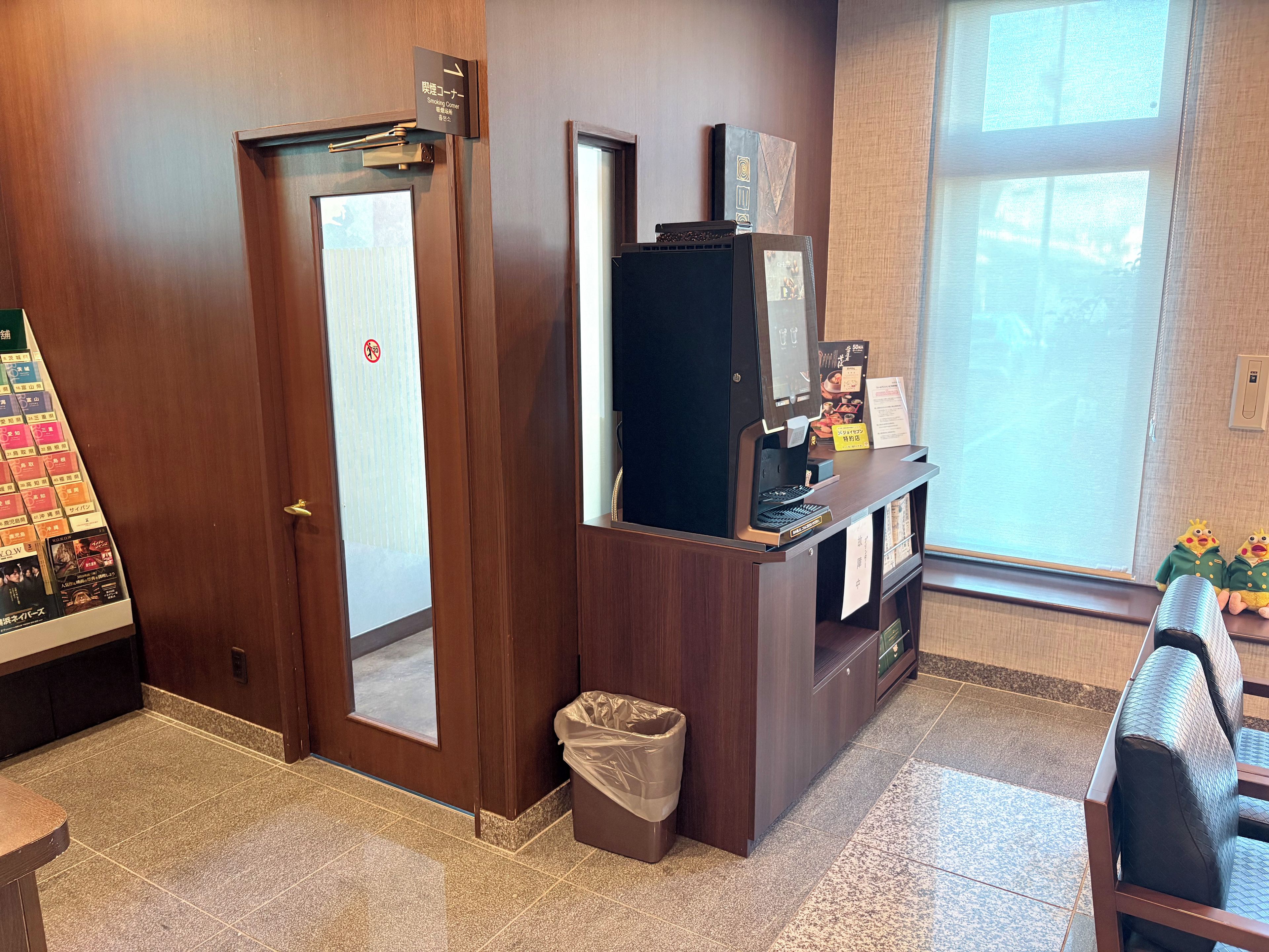 A hotel reception area with a wooden desk, a trash bin, a vending machine, and a window with blinds.