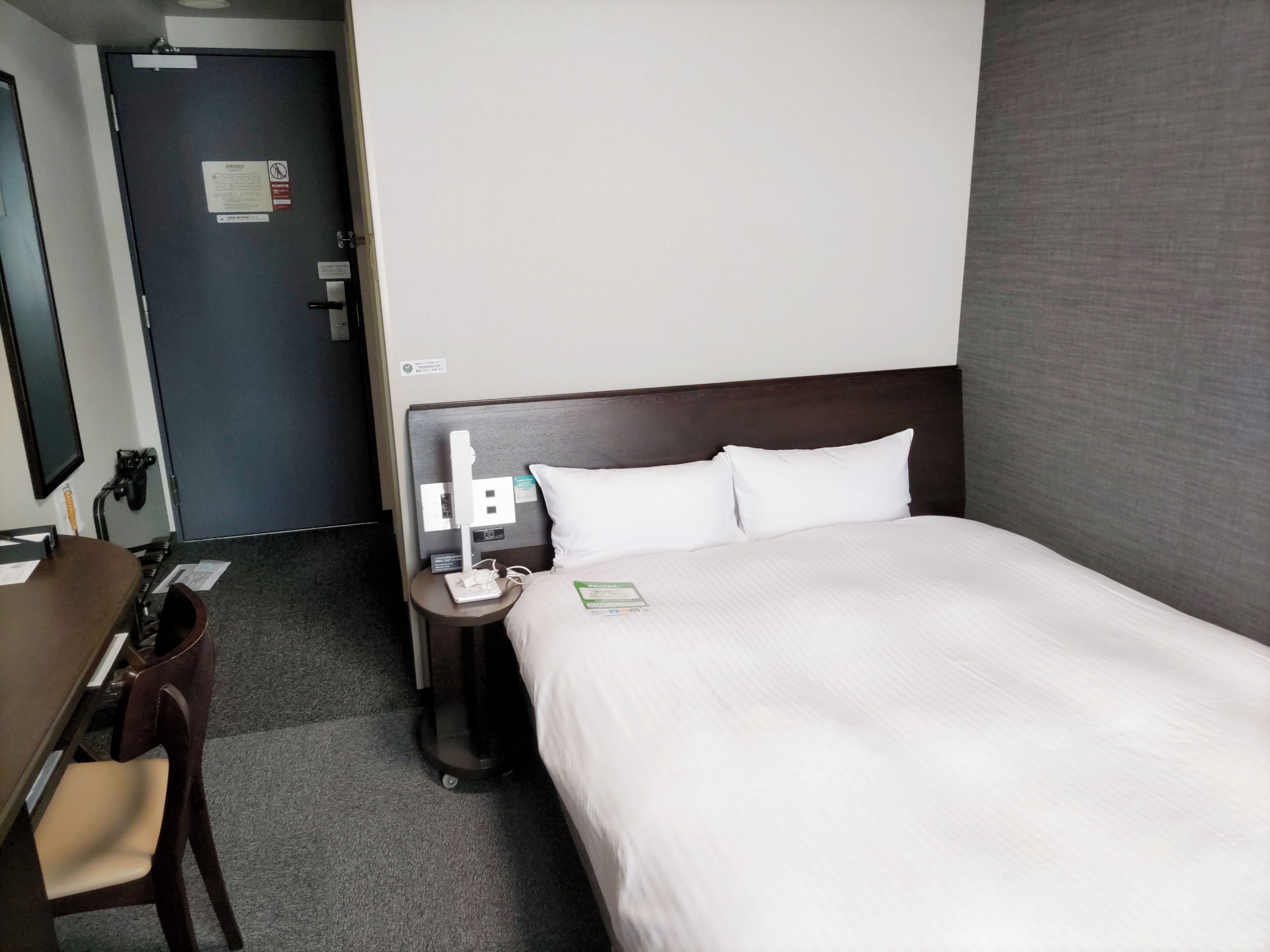 A hotel room with a bed, bedside table, lamp, chair, and a desk with a computer.