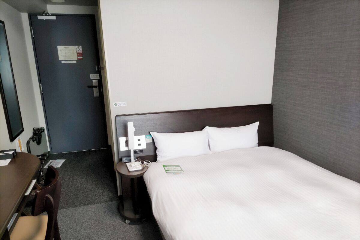 Habitació d'hotel amb un llit, una taula de nit, una cadira, un escriptori i un televisor penjat a la paret.