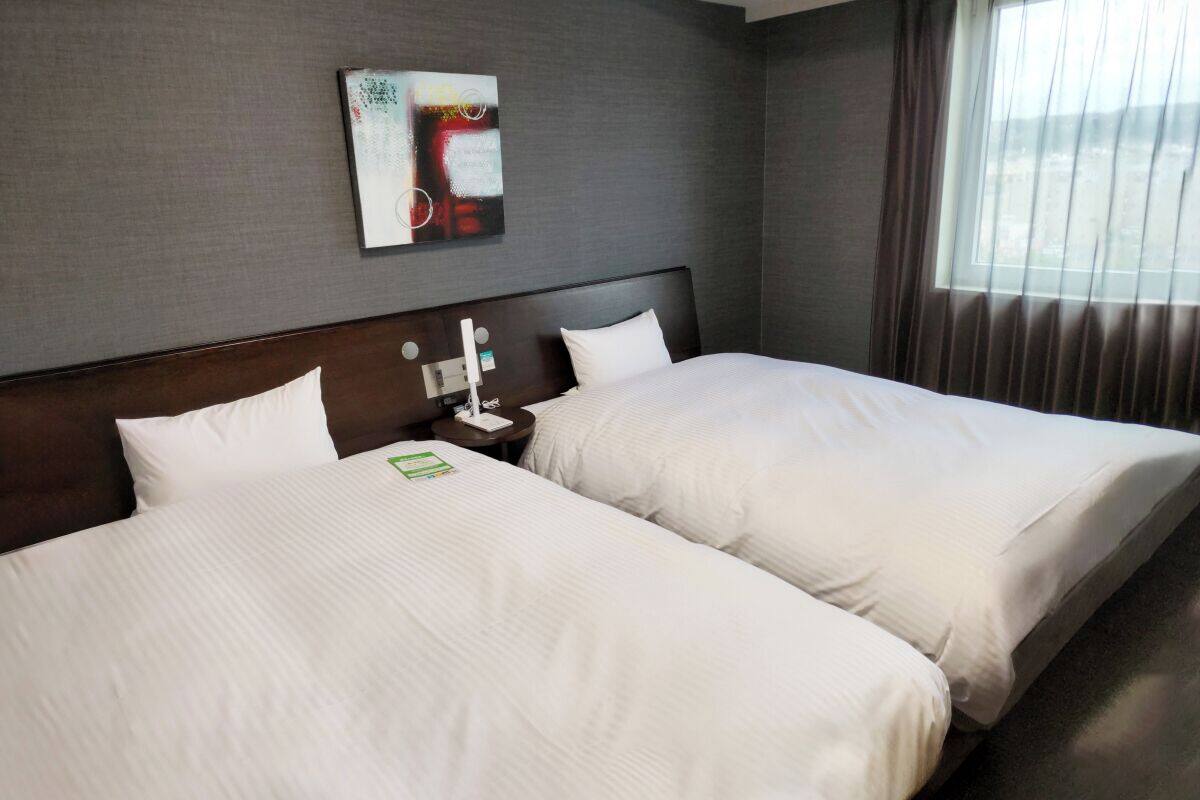 Habitació d'hotel amb dos llits, una tauleta de nit amb una làmpada, un quadre a la paret i una finestra amb cortines.
