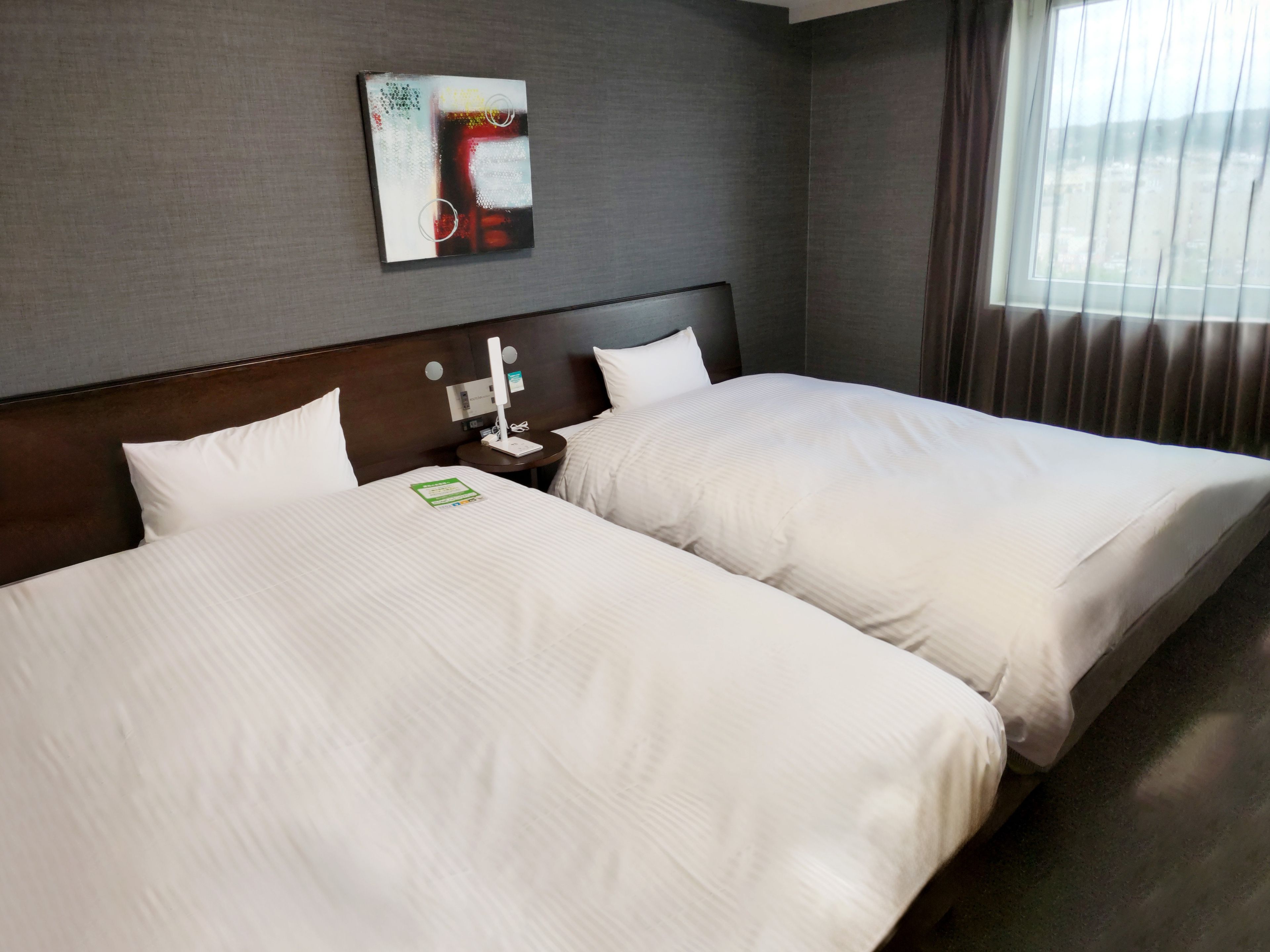 A hotel room with two beds, a nightstand with a lamp, a wall-mounted artwork, and a window with curtains.