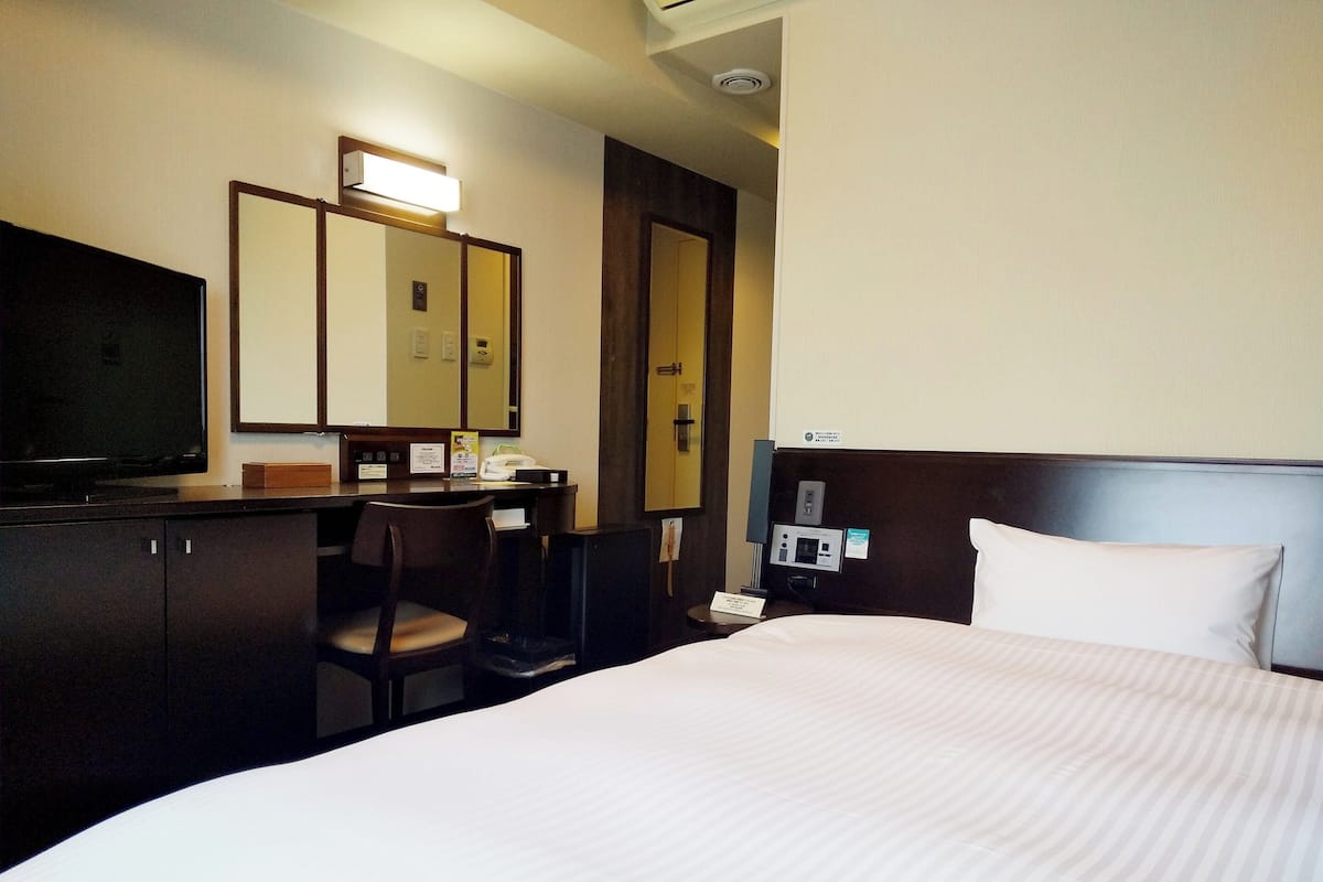 Ein Hotelzimmer mit einem Bett, einem Schreibtisch mit Stuhl, einem Fernseher, einem Spiegel und einem Nachttisch mit Telefon.