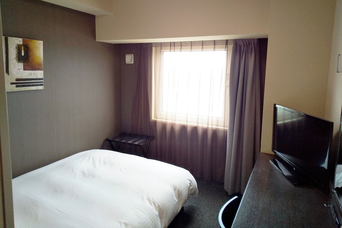 Ein Hotelzimmer mit Bett, Fernseher, kleinem Tisch, Fenster mit Vorhängen und einem wandmontierten Lichtschalter.