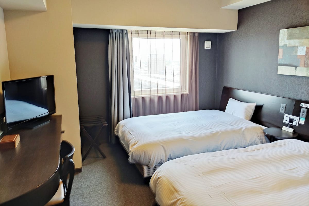 Ein Hotelzimmer mit zwei Betten, einem Schreibtisch mit Fernseher und einem Fenster mit Vorhängen.