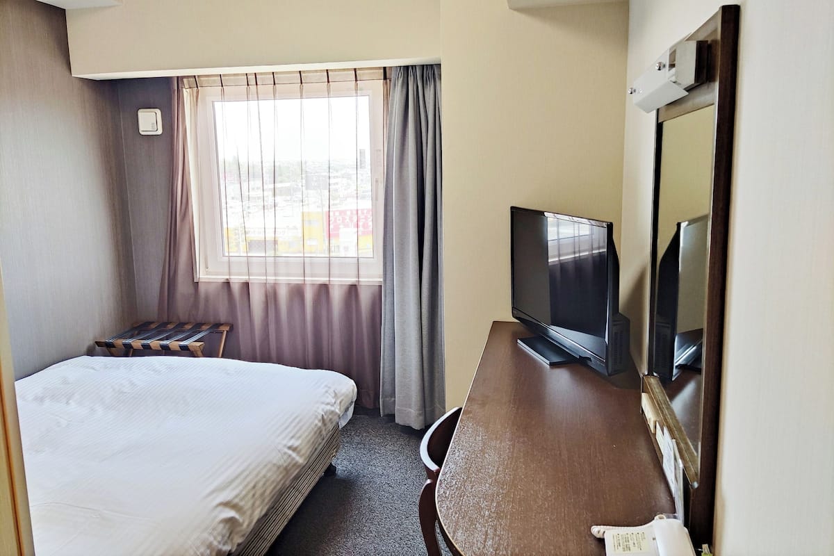 Ein Hotelzimmer mit Bett, Schreibtisch mit Fernseher, Fenster mit Vorhängen und Spiegel.
