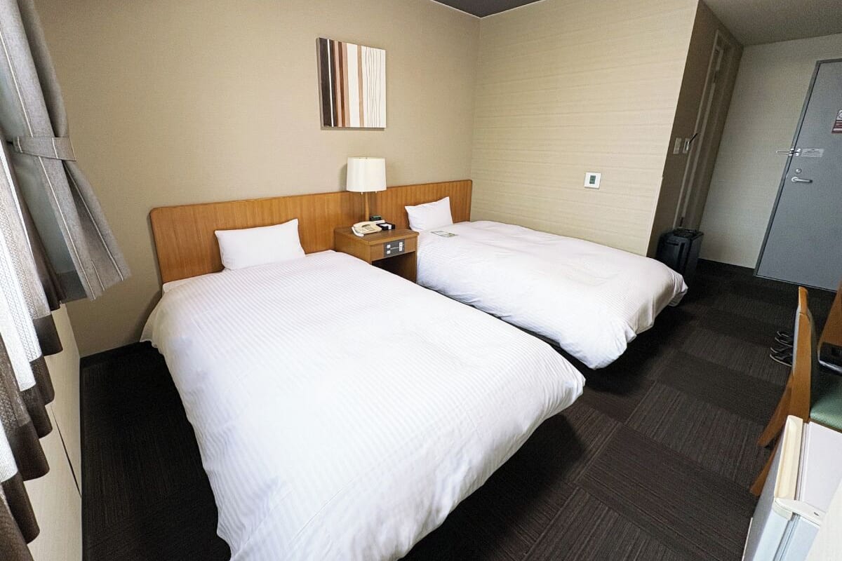 A hotel room with two beds, a wooden headboard, a bedside table with a lamp, and a small desk with a telephone.