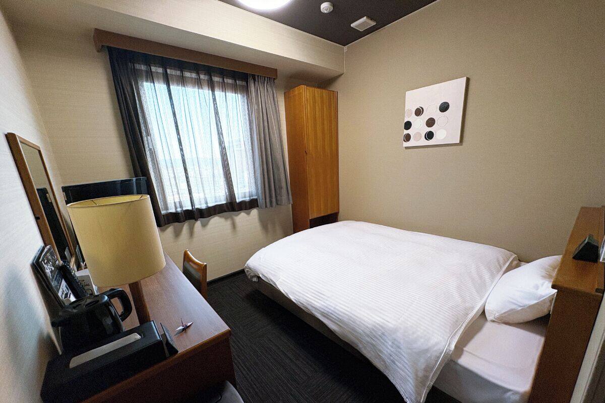 A hotel room with a bed, a desk with a lamp, a chair, a window with curtains, and a wall-mounted artwork.