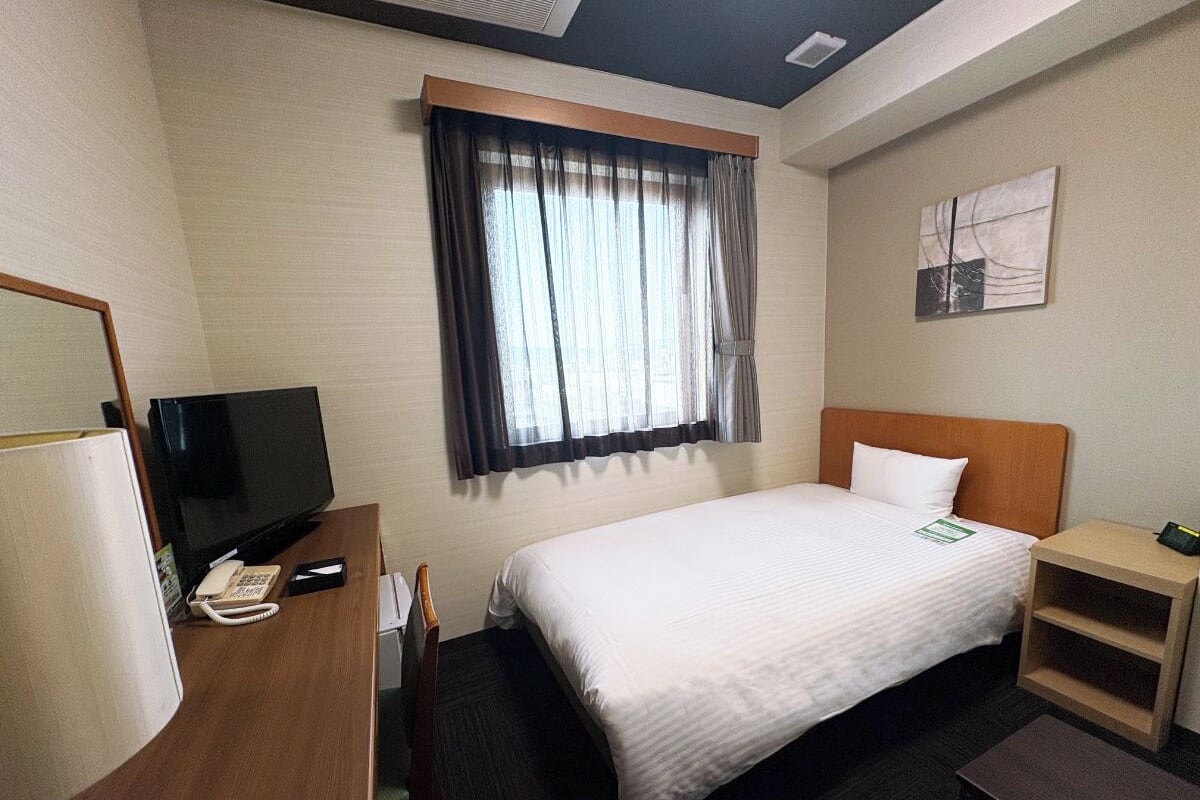 A hotel room with a bed, a desk with a telephone and a television, a window with curtains, and a small bedside table.