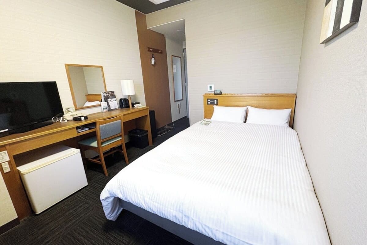 A hotel room with a bed, a desk with a computer, a television, and a small refrigerator.
