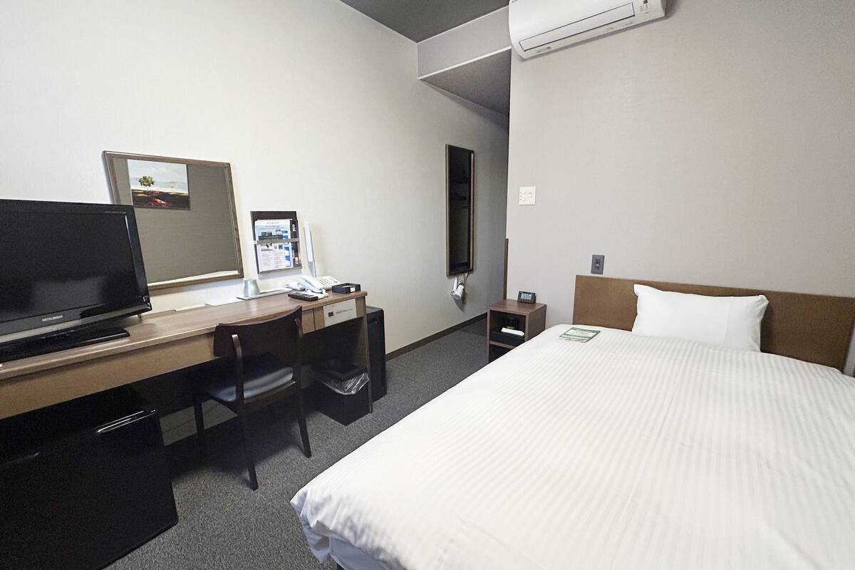 A hotel room with a bed, a desk with a computer, a chair, a small table, and an air conditioning unit.