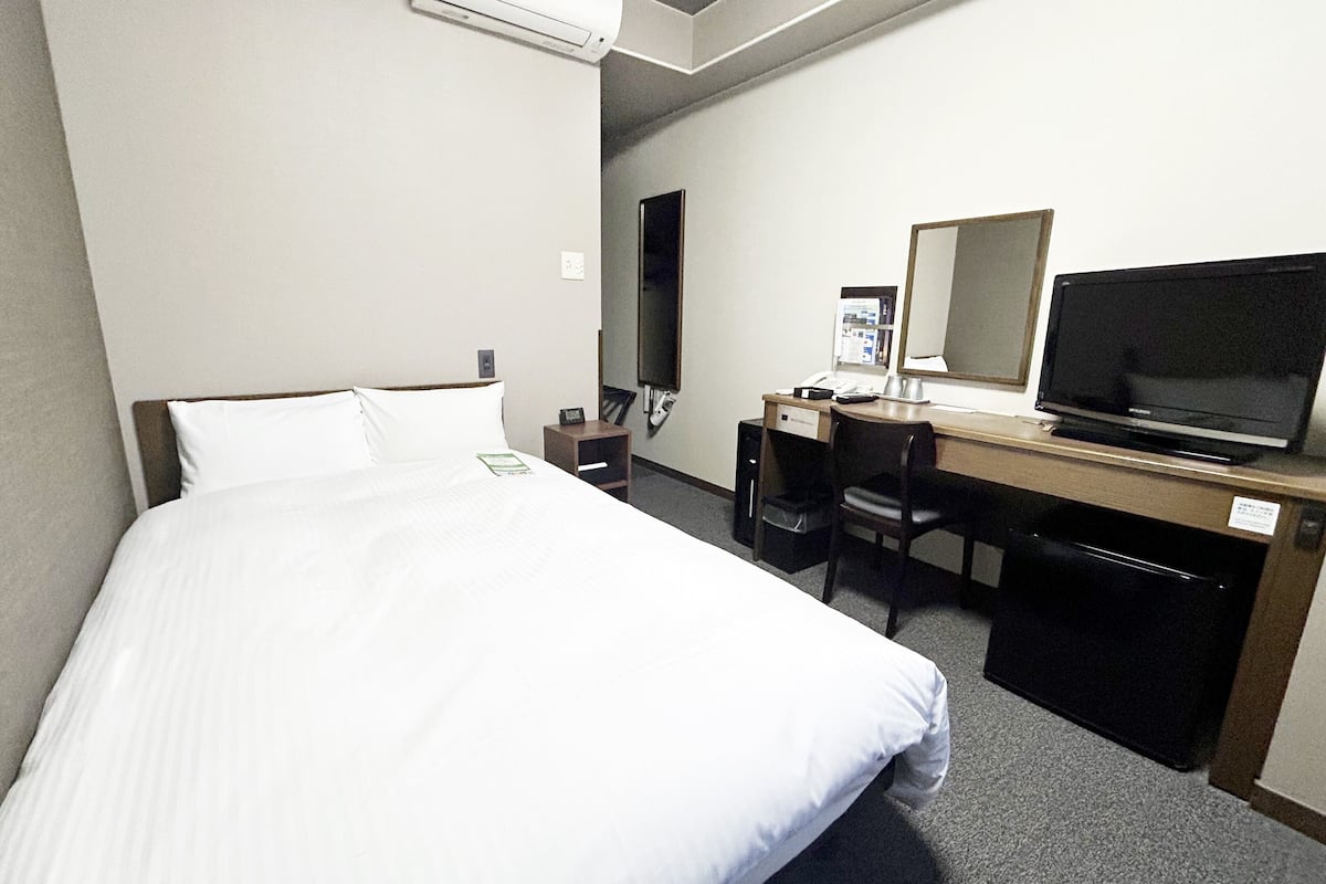 A hotel room with a bed, a desk, a chair, a television, and an air conditioning unit.