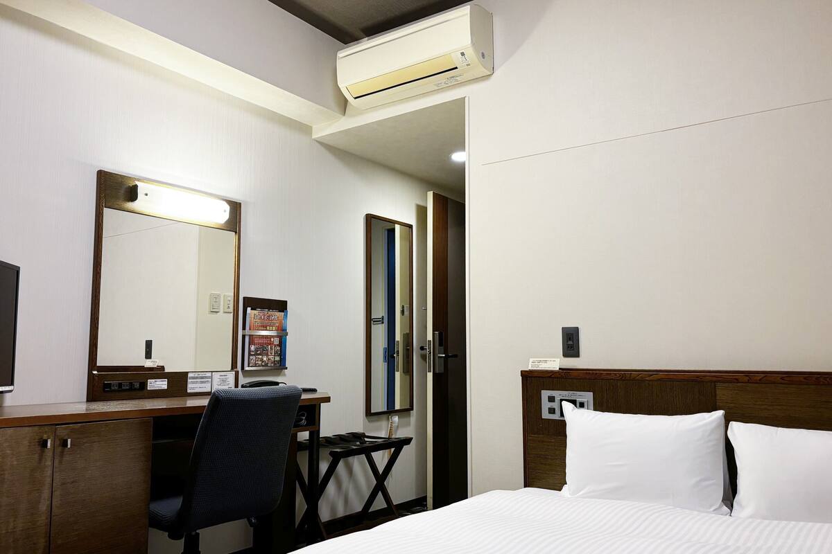 A hotel room with a bed, a desk, a chair, a mirror, and an air conditioning unit.