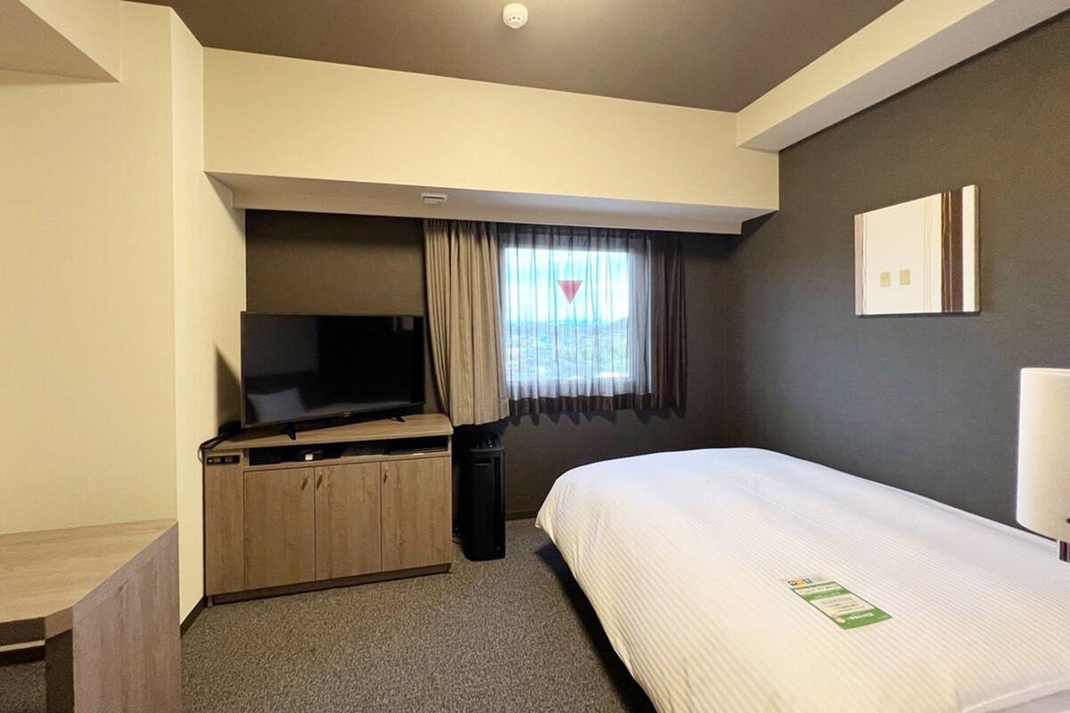 A hotel room with a bed, a TV on a wooden stand, a small desk, and a window with curtains.