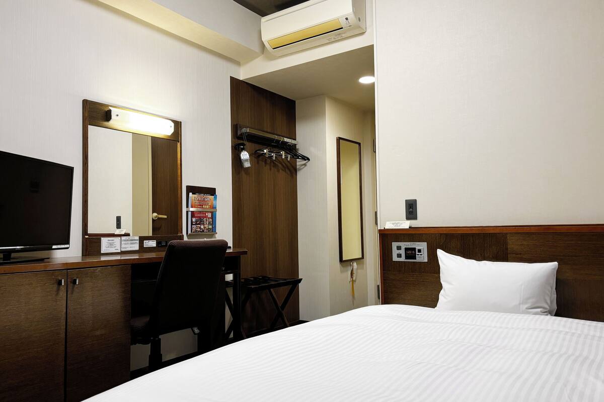 A hotel room with a bed, a desk with a computer, a television, a mirror, and an air conditioning unit.