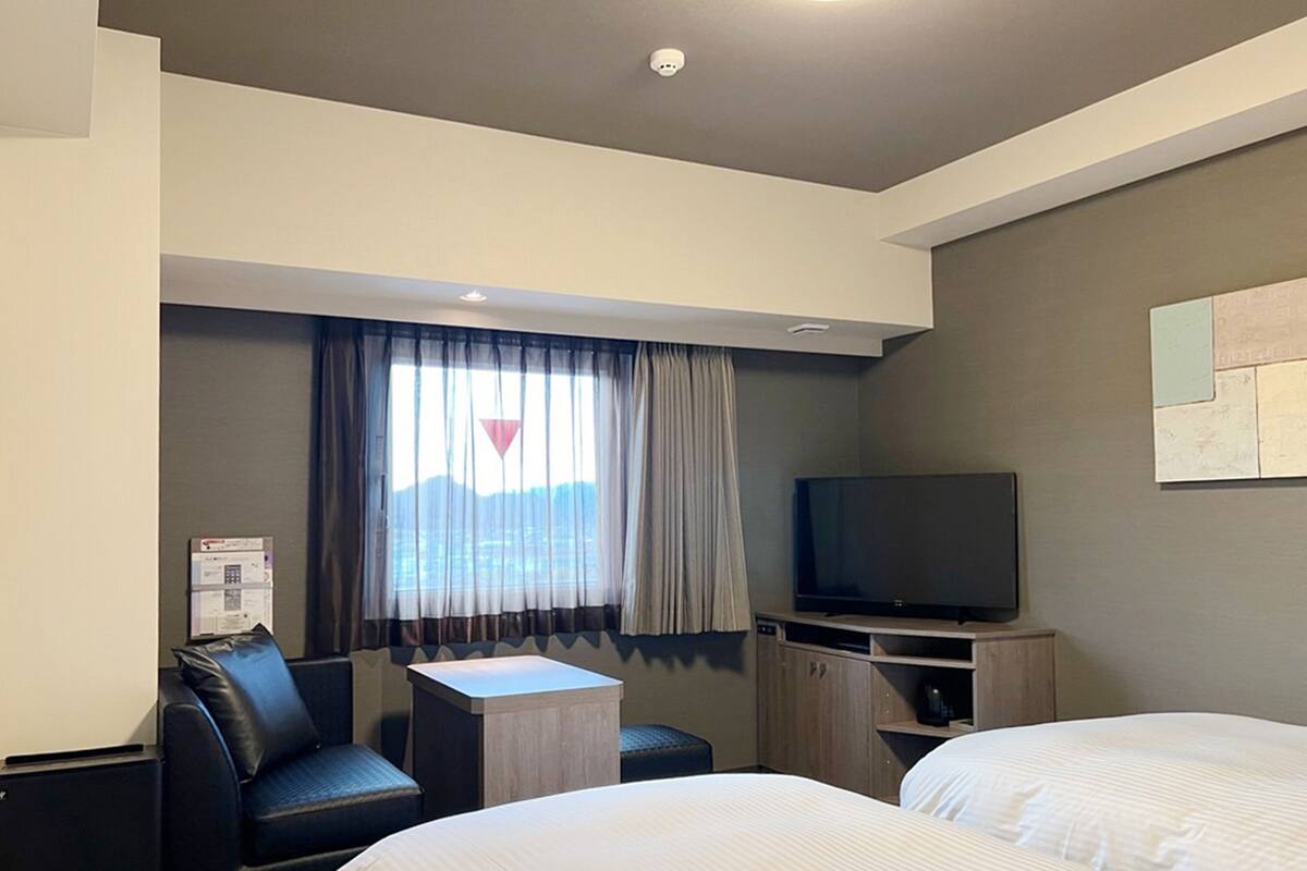 A hotel room with two beds, a TV, a small table, and a window with curtains.