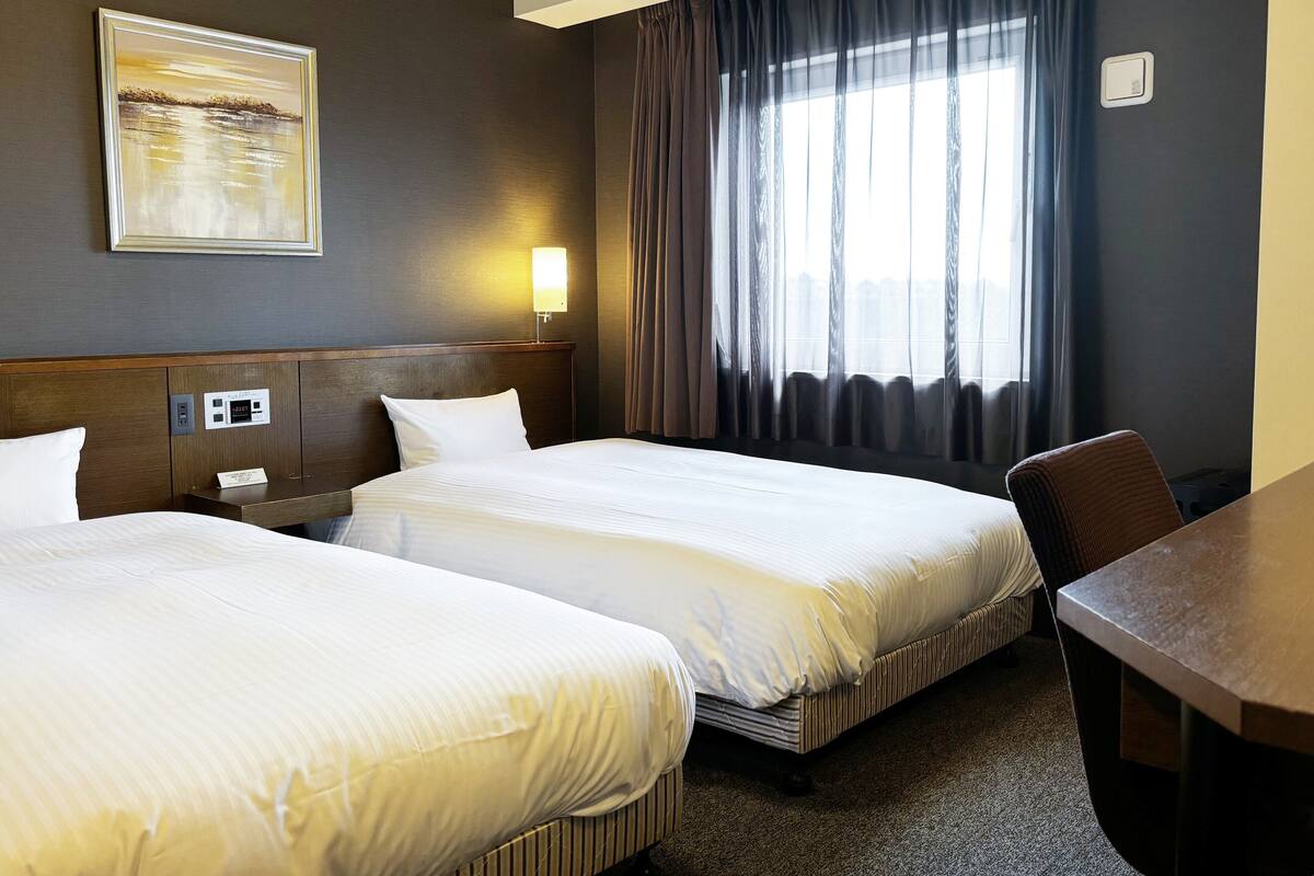 A hotel room with two beds, a desk, a nightstand, a wall clock, a painting, and a window with curtains.