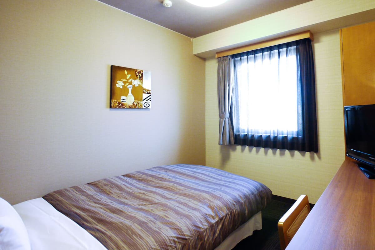 A hotel room with a single bed, a television, a window with curtains, and a painting on the wall.