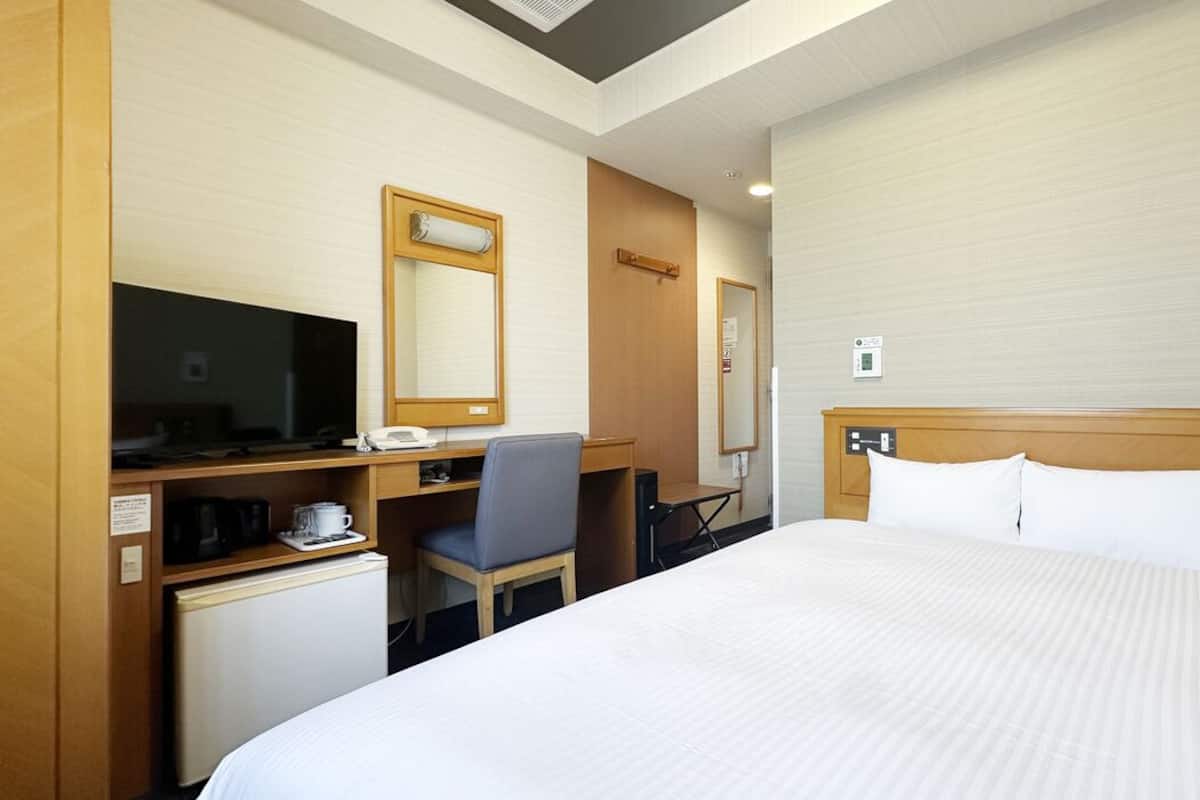 A hotel room with a bed, a desk with a computer, a chair, a small refrigerator, and a television.