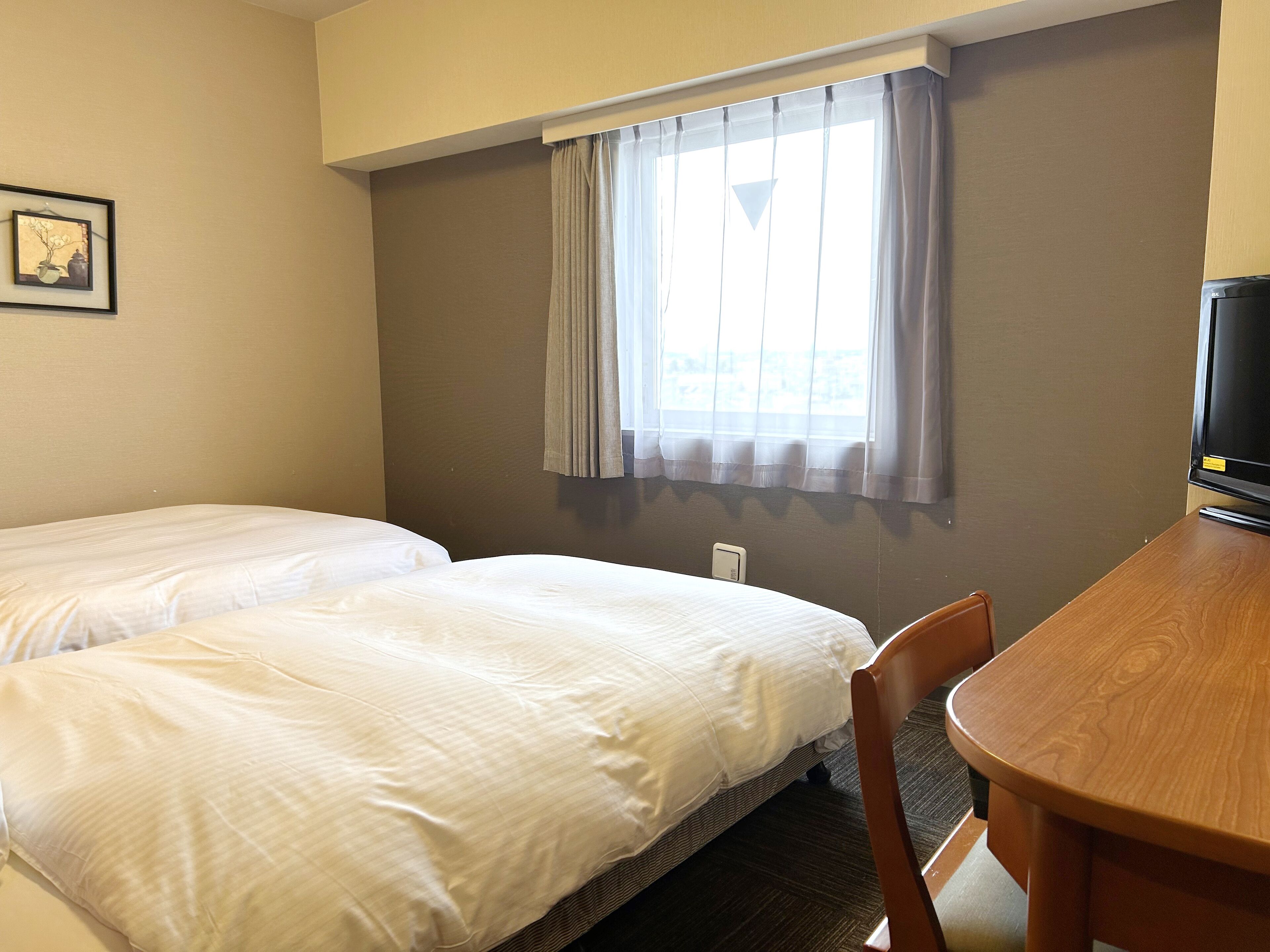 A hotel room with two beds, a wooden desk, a television, and a window with curtains.