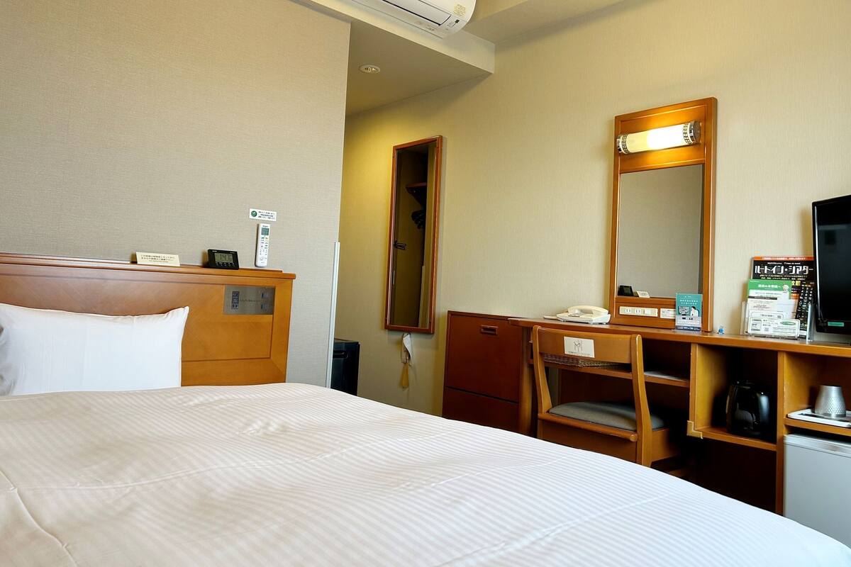 A hotel room with a bed, a desk with a telephone, a mirror, a coffee maker, and a small refrigerator.