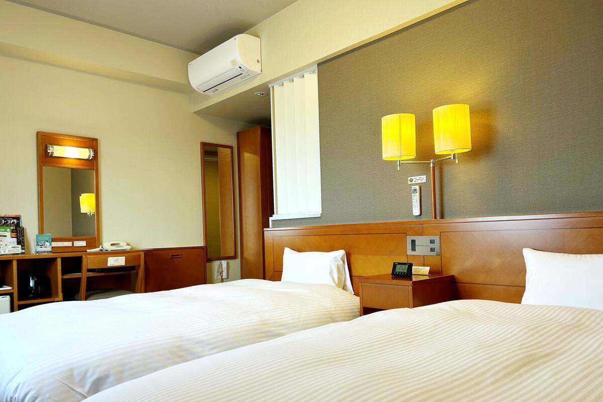 A hotel room with two beds, a desk, a mirror, a bedside table with a lamp, and an air conditioning unit.
