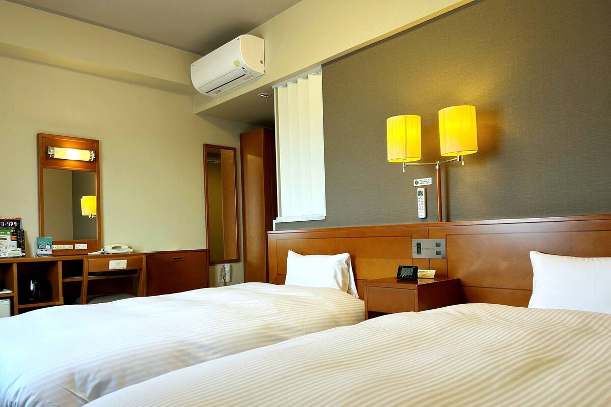 A hotel room with two beds, a desk, a mirror, and a wall-mounted air conditioner.