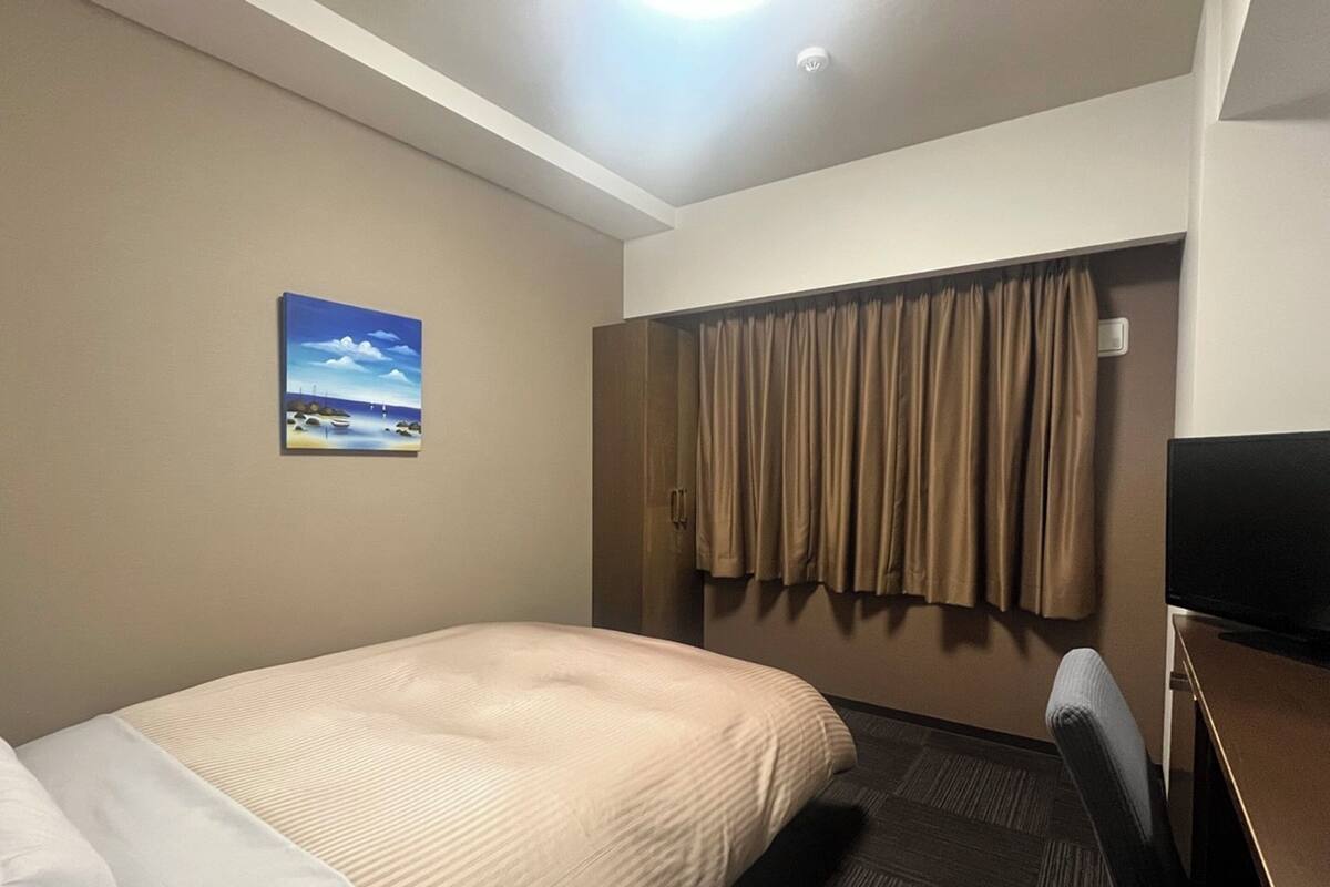 A hotel room with a bed, a desk, a television, a painting on the wall, and a window with curtains.