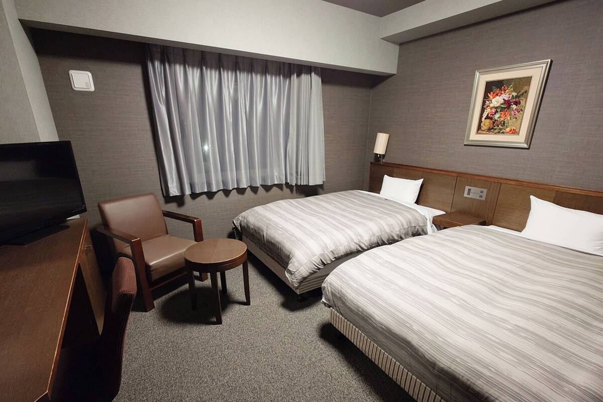 A hotel room with two beds, a desk, a chair, a small table, a TV, and a window with curtains.