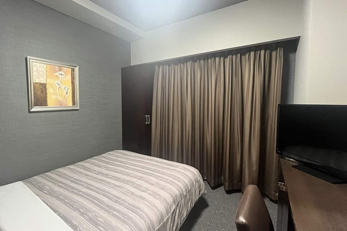 Quarto de hotel com cama, escrivaninha, cadeira, televisão, porta e cortina.