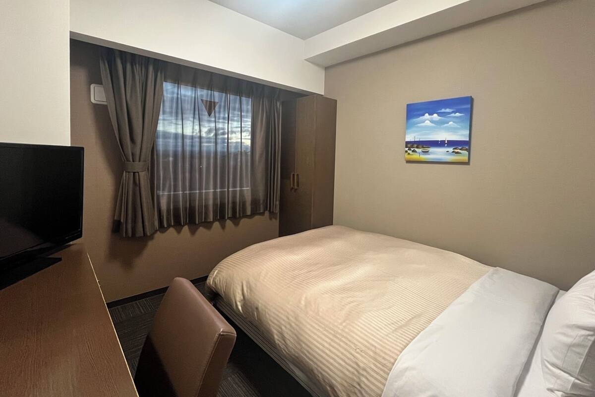 A hotel room with a bed, a TV, a window with curtains, and a painting on the wall.