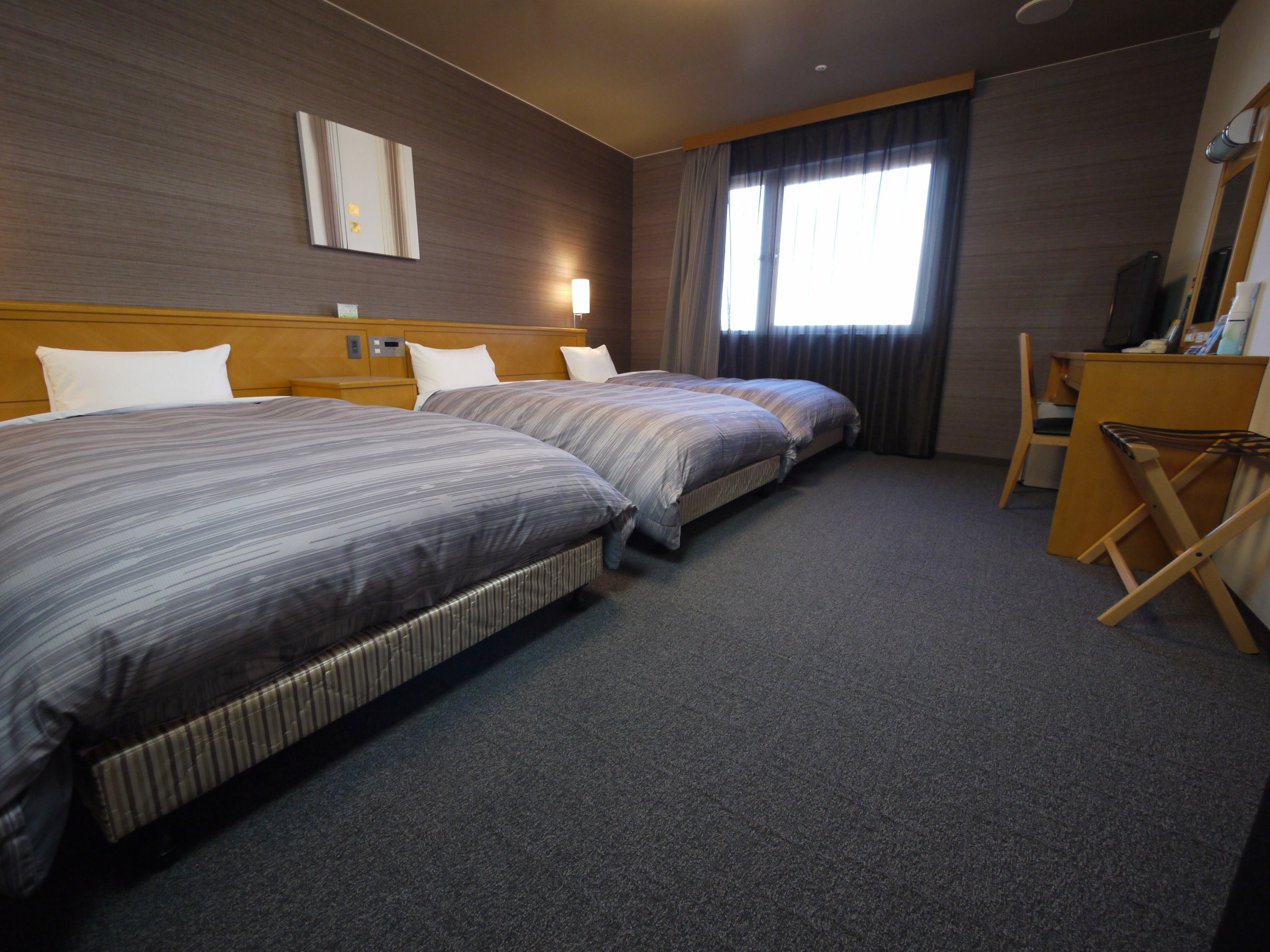 A hotel room with two beds, a desk, a chair, a radiator, and a large window with curtains.