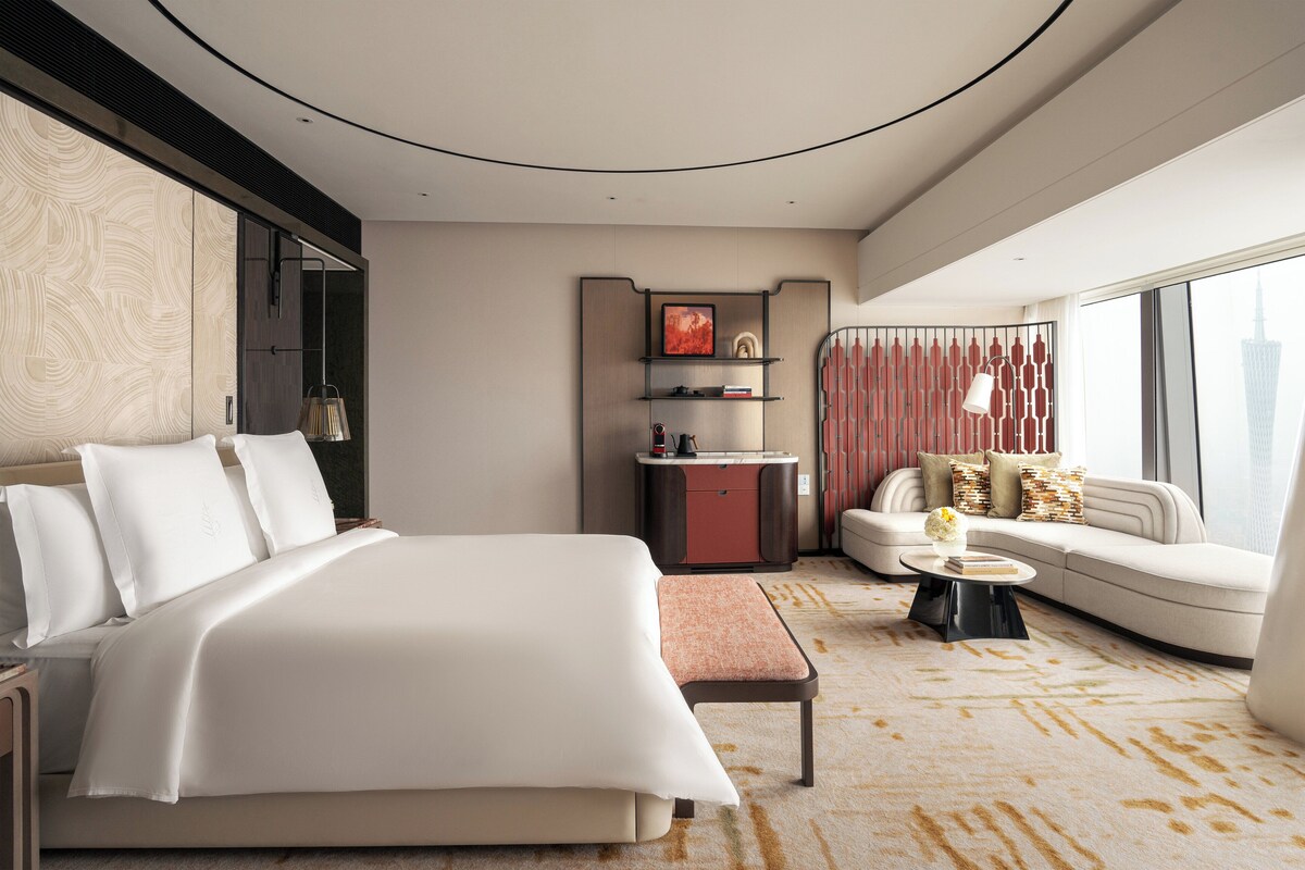 A modern hotel room with a large bed, a sofa, a small table, and a view of a cityscape.