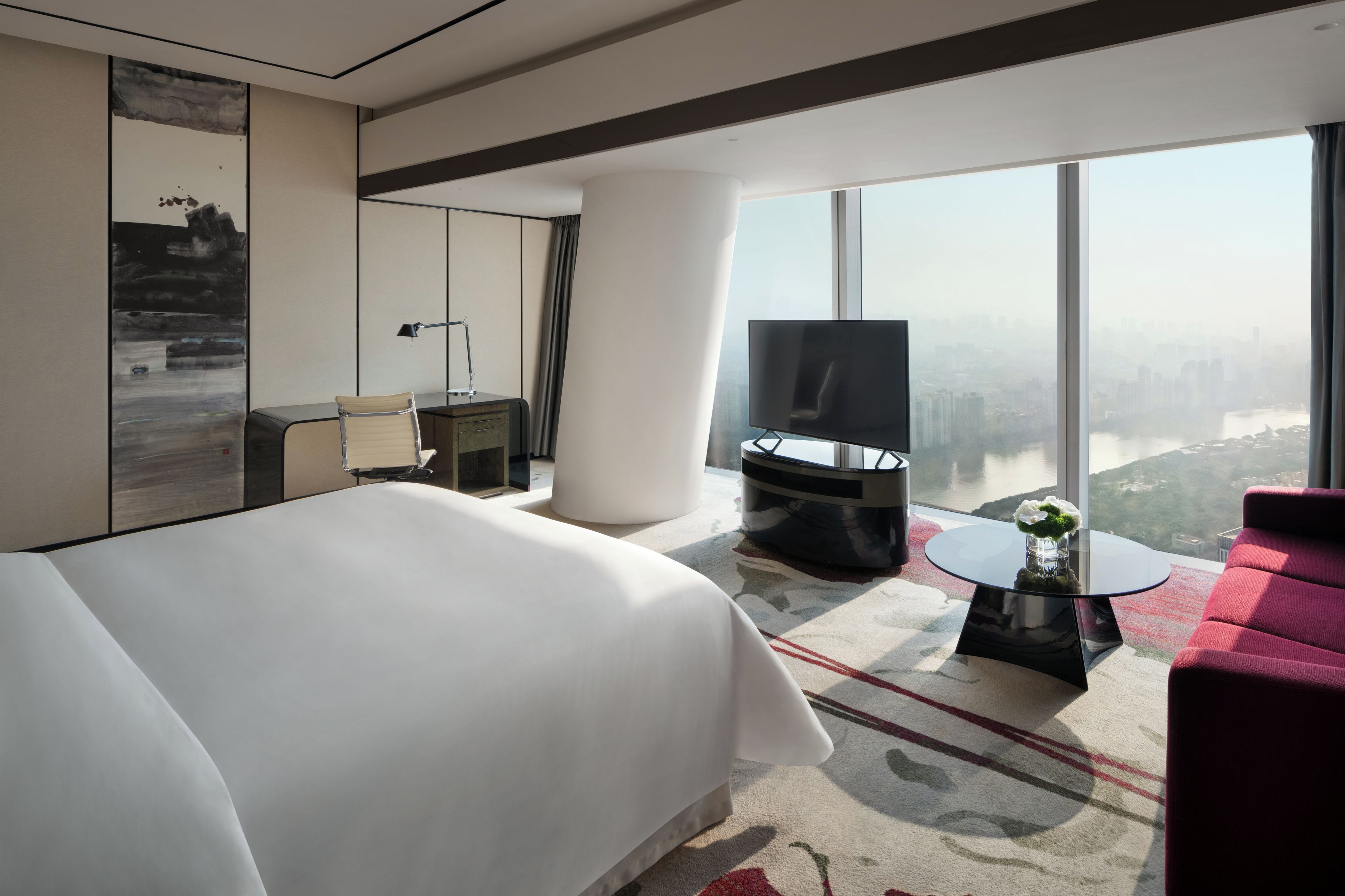 Una habitación de hotel moderna con una cama grande, televisión, sofá rojo, una pequeña mesa con un florero y vistas a la ciudad a través de amplias ventanas.