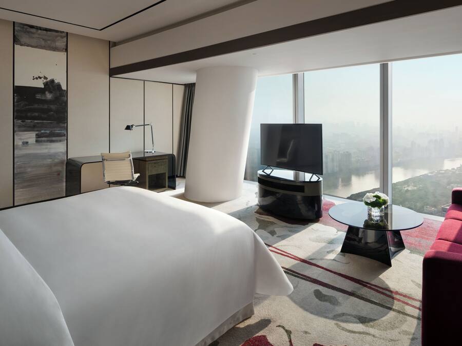 Una habitación de hotel moderna con una cama grande, televisión, sofá rojo, una pequeña mesa con un florero y vistas a la ciudad a través de amplias ventanas.