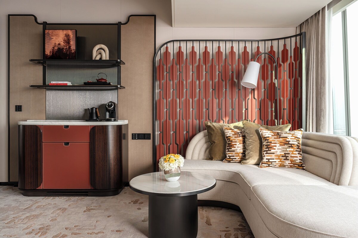 A modern hotel room with a curved sofa, a small round table with a vase, a kitchenette, and a patterned wall.