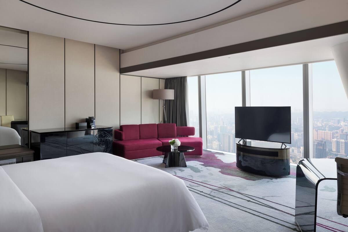 A modern hotel room with a large bed, a red sofa, a flat-screen TV, and a city view.