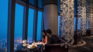 A high-rise dining area with a panoramic city view, a glass table, and a decorative chandelier.