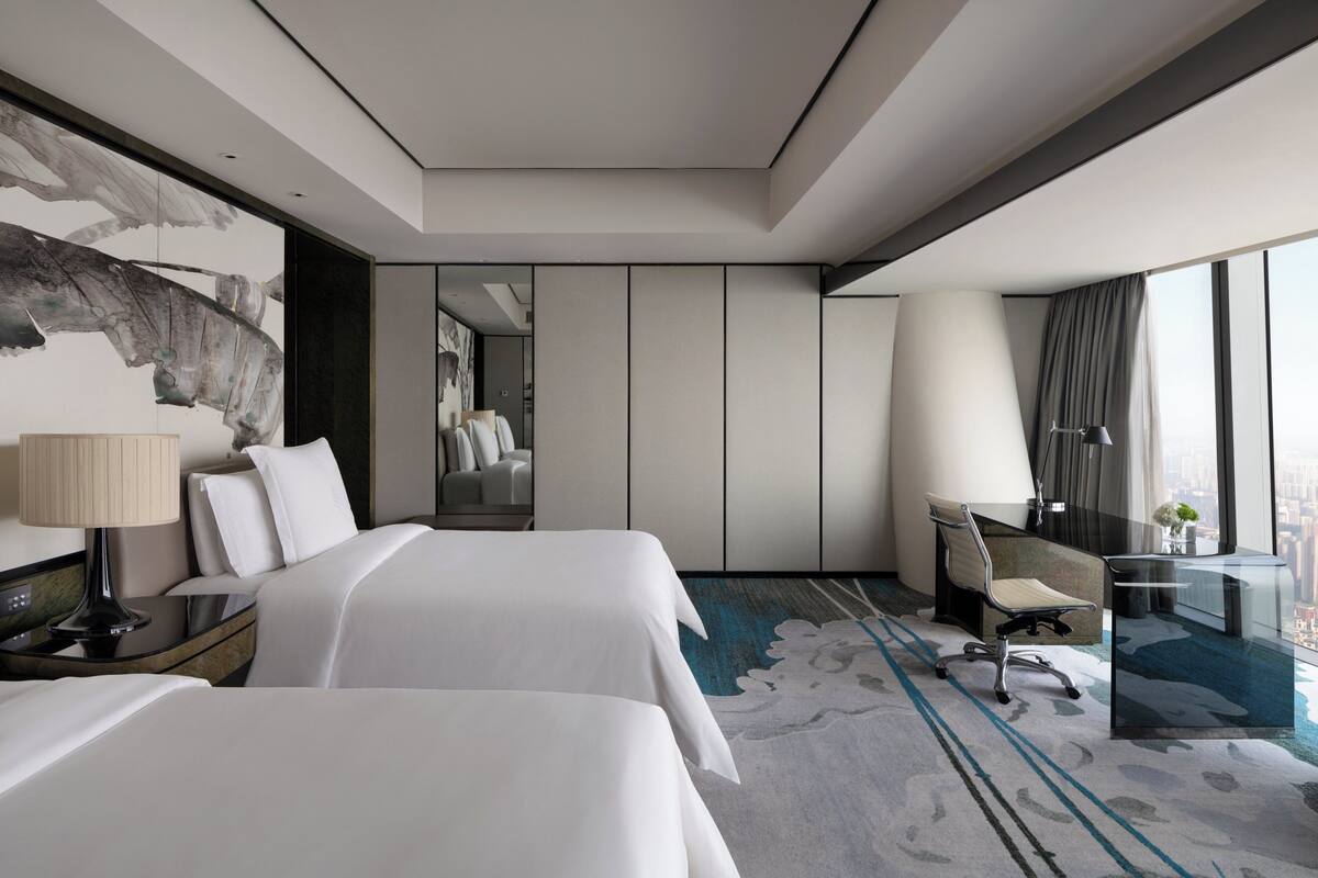 A modern hotel room with two beds, a desk, and a large window offering a city view.