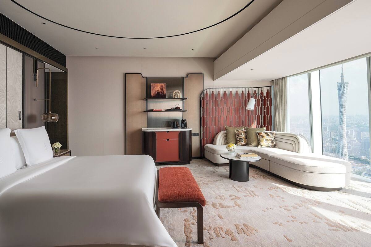 A modern hotel room with a large bed, a sofa, a small kitchenette, and a city view.