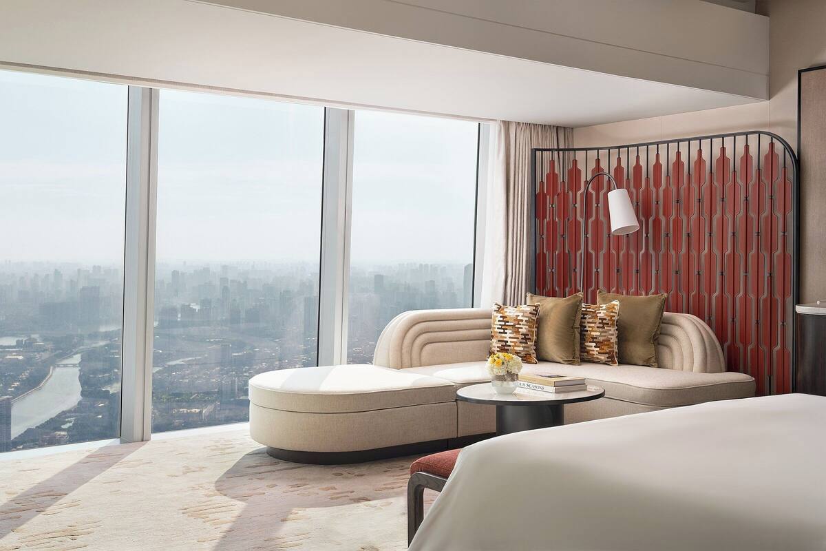 A modern hotel room with a large window offering a city view, a sofa with cushions, a small table with a vase, and a bed.