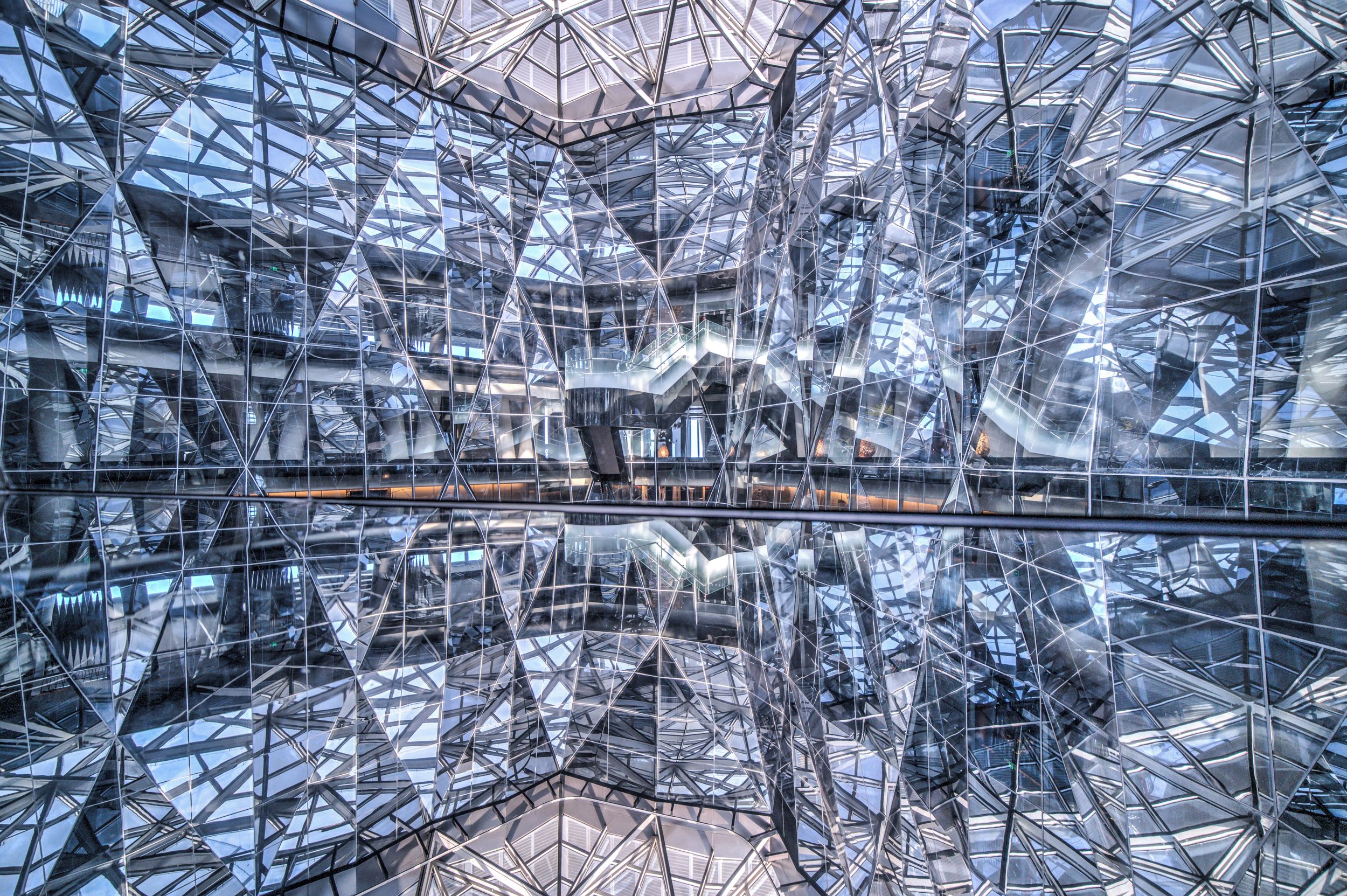 A modern architectural structure with a complex geometric design, featuring a reflective glass facade and a central open space.