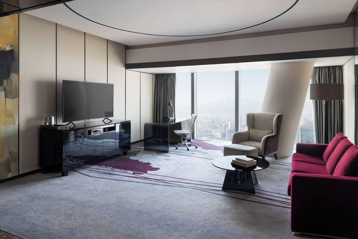 A modern hotel room with a large window, a flat-screen TV, a desk, a chair, a sofa, and a coffee table.