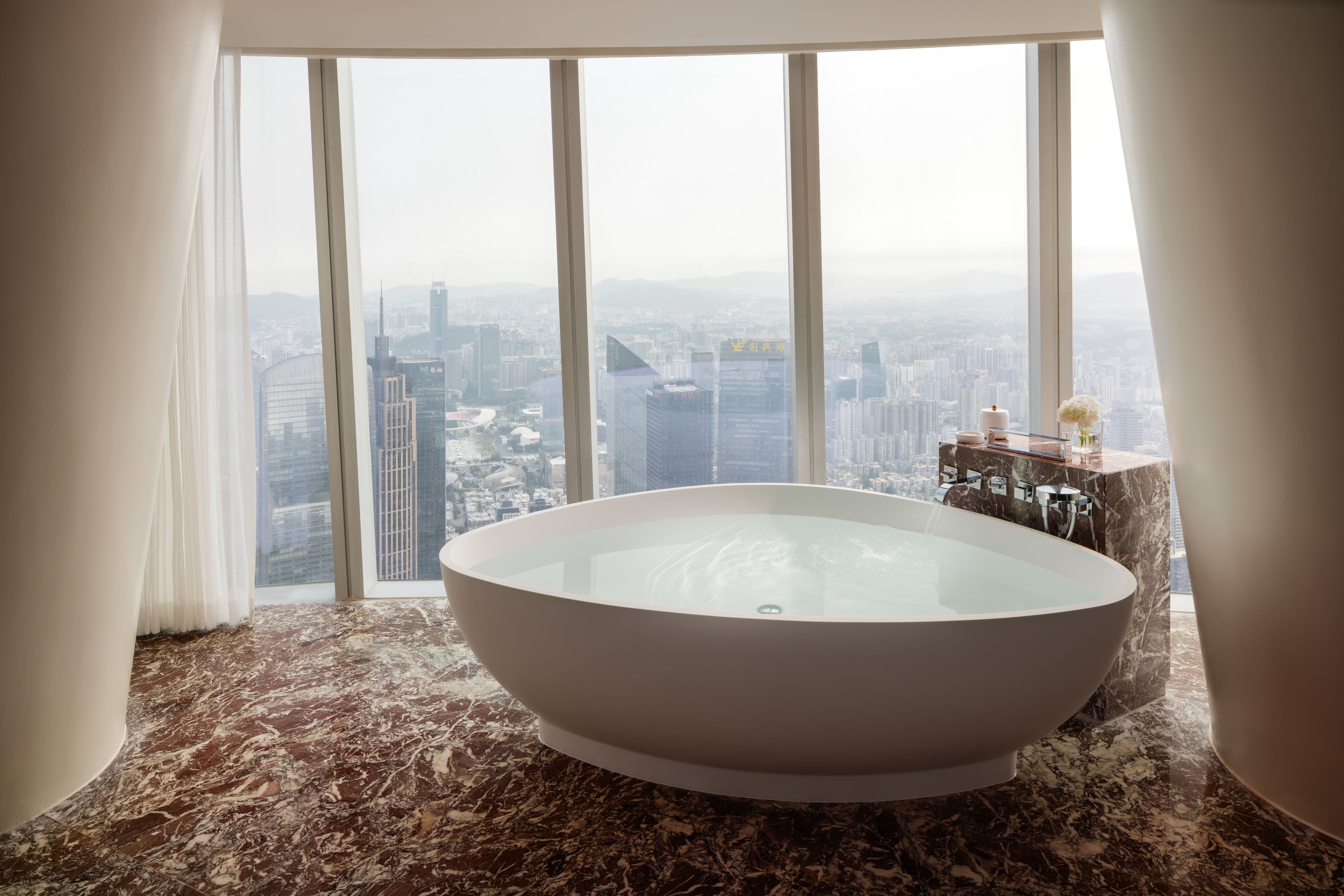 Une salle de bain moderne avec une grande baignoire sur pieds, un revêtement de sol en marbre et une vue sur la ville grâce à de grandes fenêtres du sol au plafond.