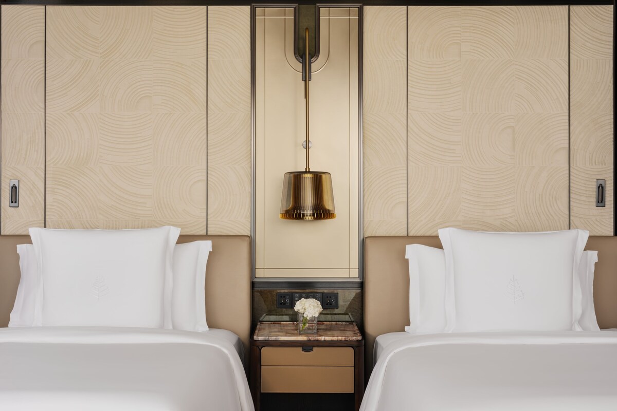 A hotel room with two beds, a nightstand with a vase of flowers, and a wall-mounted lamp.