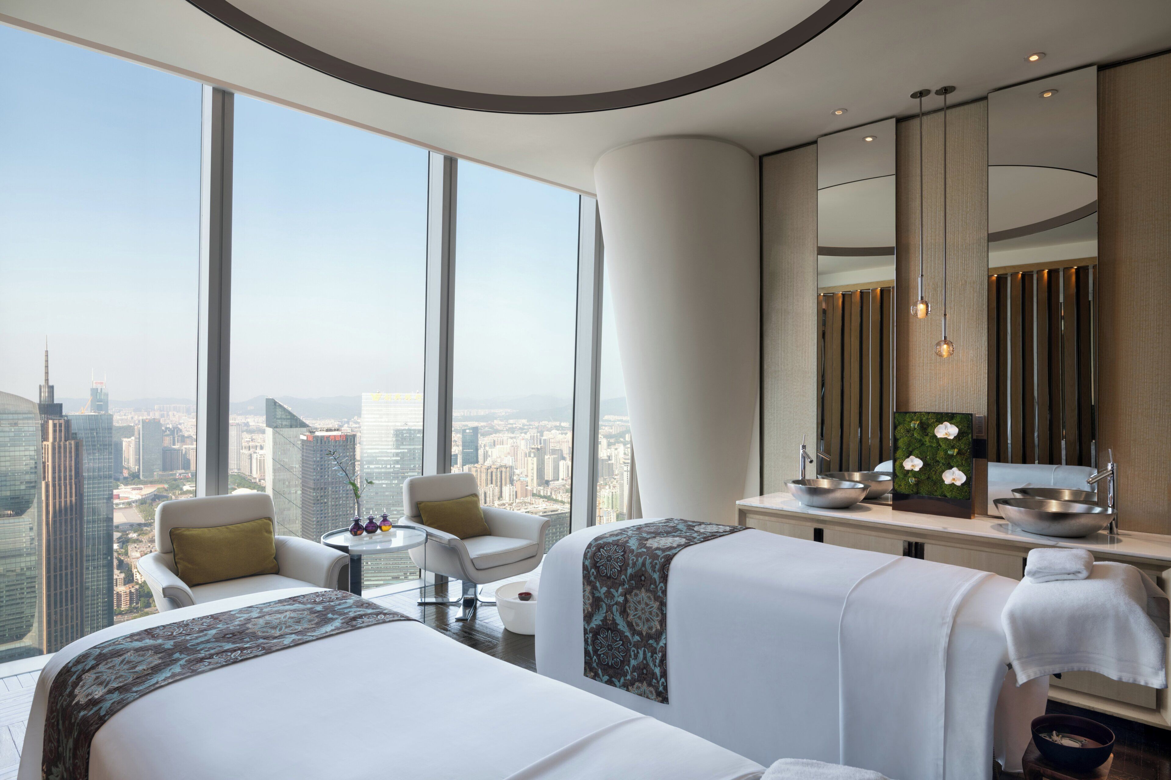 A high-rise hotel room with a city view, two beds, a sitting area, and a vanity with a mirror and sink.
